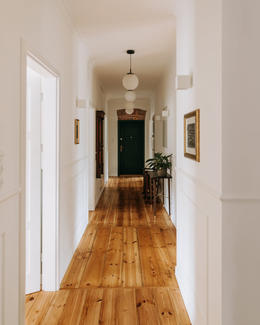 Długi korytarz z drewnianą podłogą  oraz sztukaterią na białej ścianie