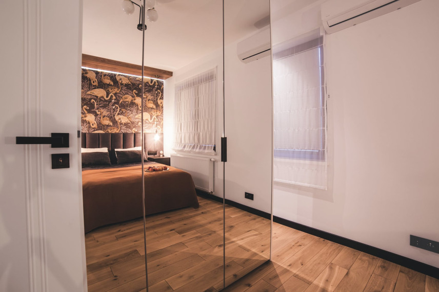 Sypialnia z tapicerowanym łóżkiem oraz szafą w zabudowie z lustrzanym frontem i ciemną podłogą