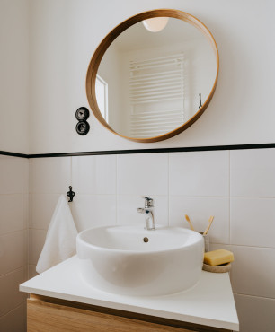 Łazienka z szafką z drewnianym frontem oraz z okrągłym lustrem w drewnianej ramie umieszczone na białej ścianie