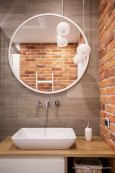 Mała łazienka z cegłą na ścianie oraz szarymi płytkami oraz okrągłym lustrem zamontowanym nad umywalką