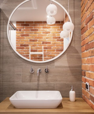 Mała łazienka z cegłą na ścianie oraz szarymi płytkami oraz okrągłym lustrem zamontowanym nad umywalką